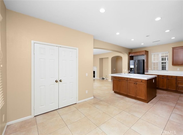 kitchen featuring black fridge with ice dispenser, arched walkways, an island with sink, light countertops, and light tile patterned floors
