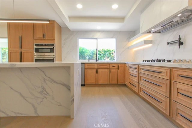 kitchen featuring custom exhaust hood, appliances with stainless steel finishes, light wood-style floors, a raised ceiling, and light stone counters