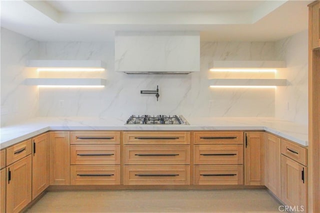 kitchen featuring stainless steel gas cooktop, ventilation hood, backsplash, a tray ceiling, and light stone counters