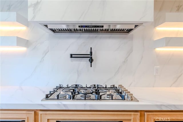 kitchen view of stainless steel gas cooktop, extractor fan, pot filler, backsplash, and light brown cabinetry