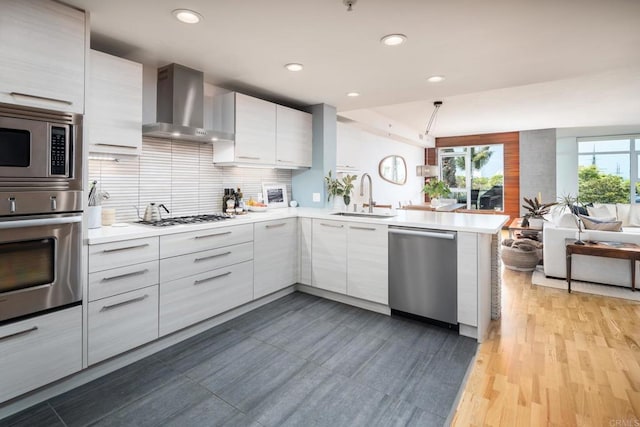 kitchen with stainless steel appliances, wall chimney exhaust hood, a sink, open floor plan, and a peninsula