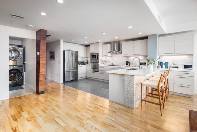 kitchen with stainless steel appliances, wall chimney range hood, stacked washer / drying machine, modern cabinets, and a sink