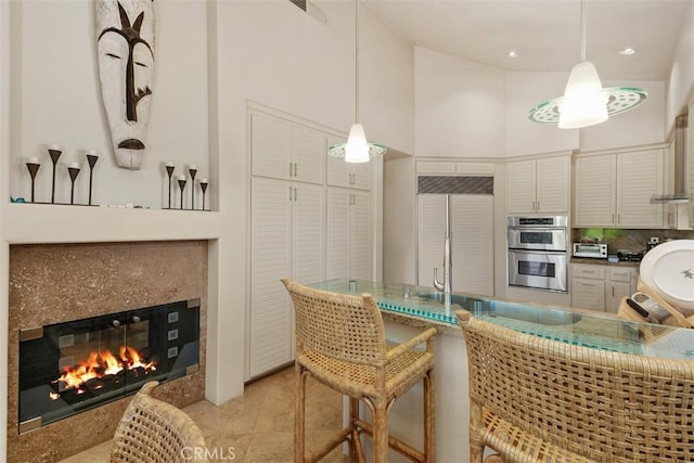 kitchen featuring stainless steel double oven, backsplash, hanging light fixtures, a high end fireplace, and a towering ceiling