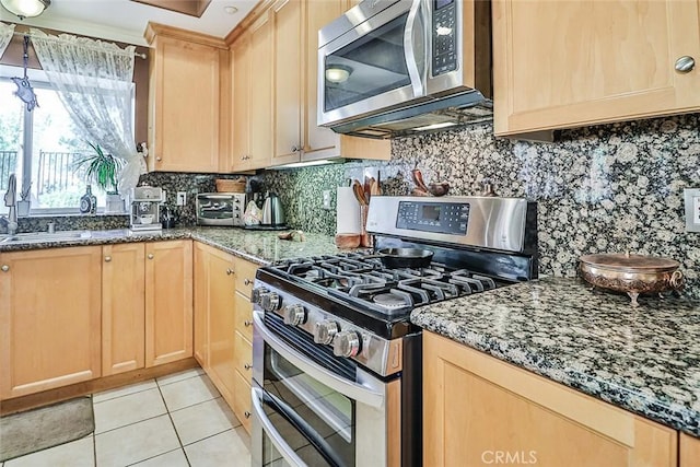 kitchen featuring stainless steel appliances, a sink, light brown cabinetry, decorative backsplash, and dark stone countertops