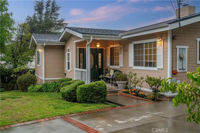 exterior entry at dusk with covered porch, a yard, and a shingled roof