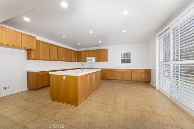 kitchen with white microwave, light countertops, a kitchen island with sink, recessed lighting, and brown cabinetry