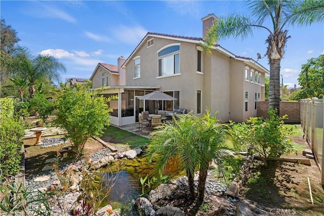 back of house featuring stucco siding, a sunroom, a chimney, a small pond, and fence