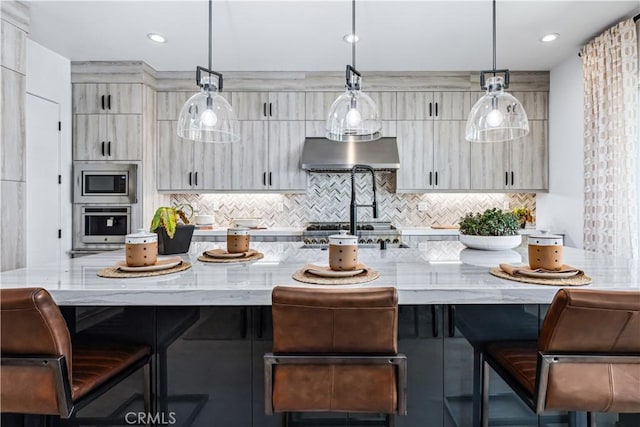 kitchen featuring appliances with stainless steel finishes, light stone countertops, a large island, under cabinet range hood, and decorative backsplash