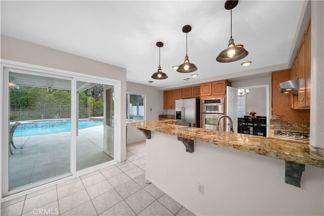 kitchen with stainless steel appliances, a peninsula, brown cabinets, pendant lighting, and wall chimney exhaust hood