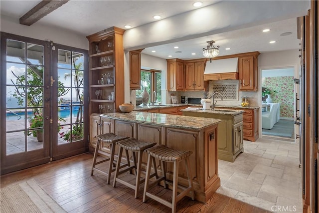 kitchen featuring custom range hood, a center island with sink, a sink, french doors, and light stone countertops