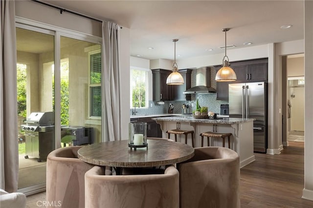 kitchen featuring wall chimney range hood, high end refrigerator, a center island, tasteful backsplash, and dark wood-style flooring