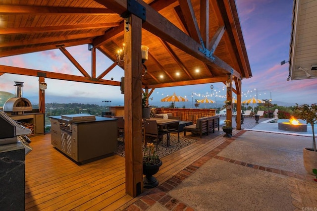 deck at dusk featuring an outdoor fire pit, a gazebo, an outdoor kitchen, and grilling area