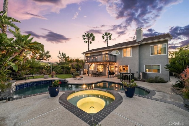 back of property at dusk with a chimney, a patio, stucco siding, a balcony, and a pool with connected hot tub