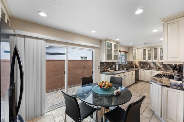 kitchen featuring cream cabinets, stone tile flooring, dishwashing machine, backsplash, and glass insert cabinets