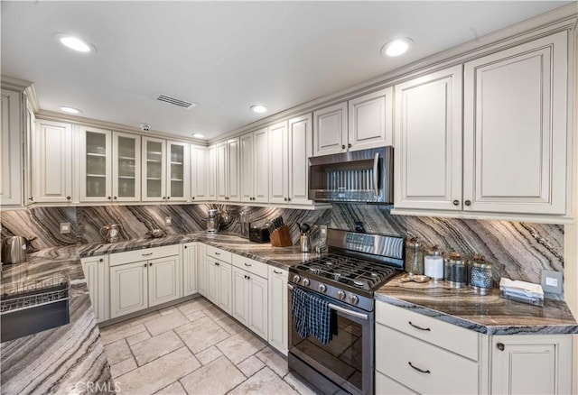 kitchen featuring appliances with stainless steel finishes, tasteful backsplash, glass insert cabinets, dark stone countertops, and light stone finish flooring