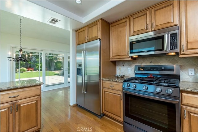kitchen with appliances with stainless steel finishes, a chandelier, light wood-type flooring, backsplash, and light stone counters