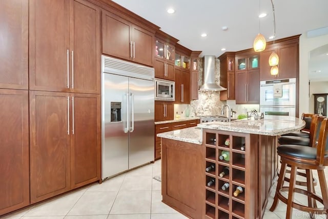 kitchen with built in appliances, wall chimney exhaust hood, tasteful backsplash, light stone counters, and pendant lighting