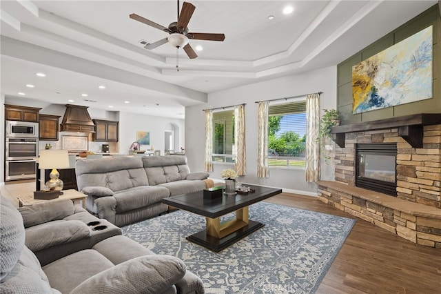 living room featuring a ceiling fan, recessed lighting, a stone fireplace, a tray ceiling, and wood finished floors