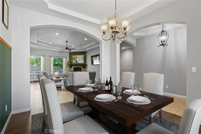 tiled dining space with a tray ceiling, arched walkways, ceiling fan, a fireplace, and a chandelier