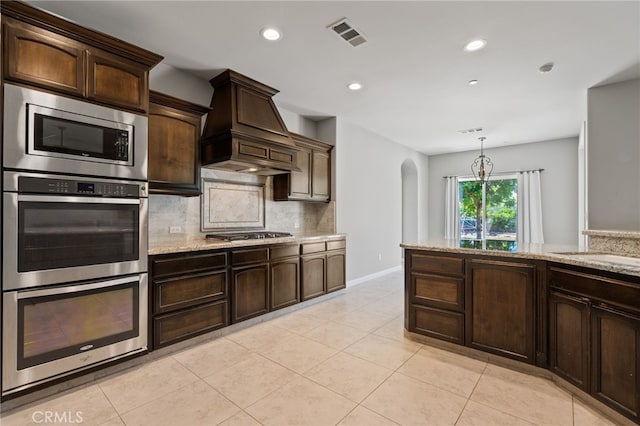 kitchen featuring appliances with stainless steel finishes, premium range hood, decorative backsplash, dark brown cabinets, and light stone counters
