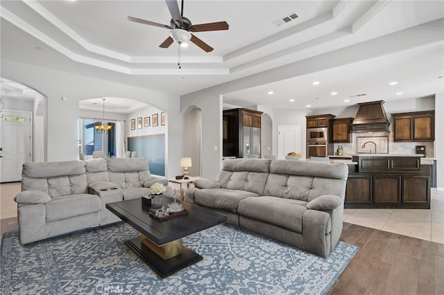 living area featuring arched walkways, a raised ceiling, ceiling fan, light wood finished floors, and a chandelier
