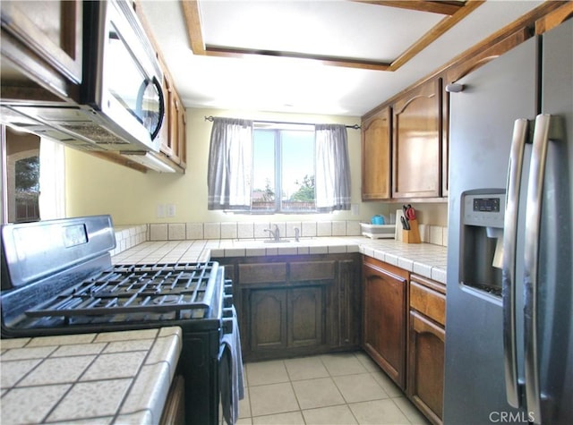 kitchen with stainless steel appliances, tile countertops, light tile patterned flooring, a sink, and brown cabinetry