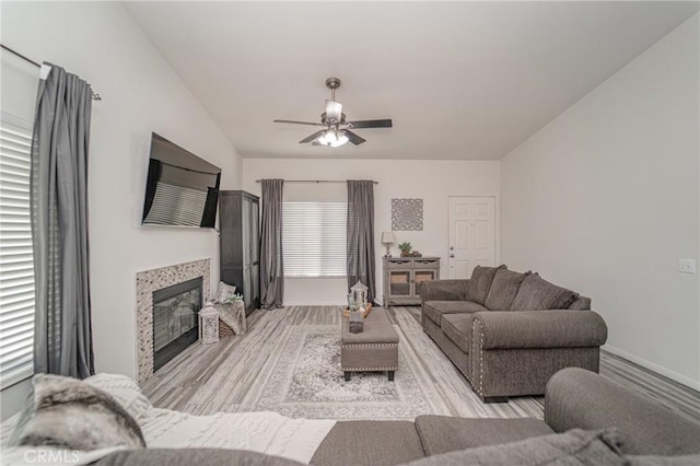 living room with light wood-type flooring, a ceiling fan, a glass covered fireplace, and baseboards