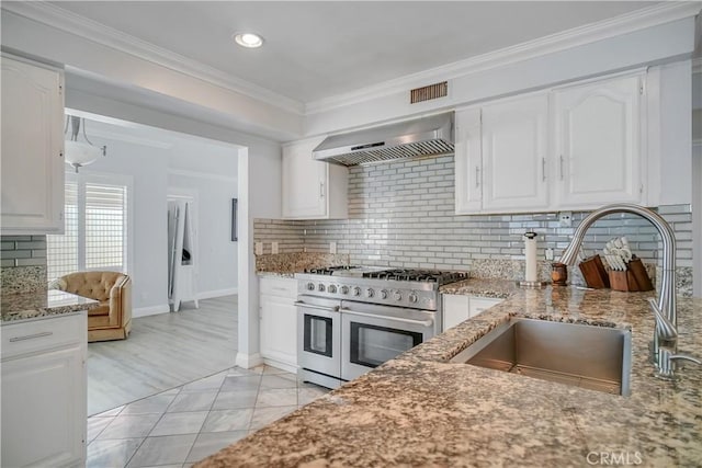 kitchen featuring double oven range, wall chimney exhaust hood, a sink, light stone counters, and white cabinets