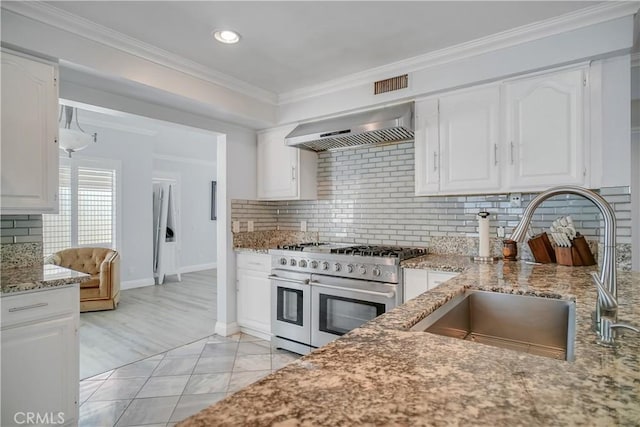 kitchen featuring double oven range, a sink, wall chimney range hood, light stone counters, and white cabinets