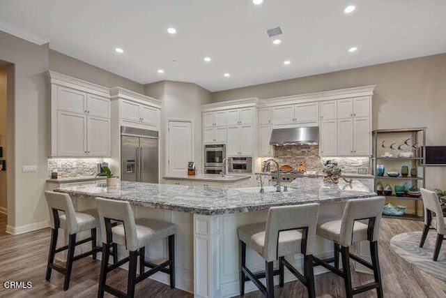 kitchen with built in refrigerator, under cabinet range hood, a sink, dark wood-style flooring, and a large island
