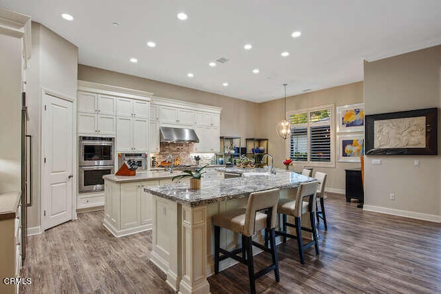 kitchen featuring a large island, under cabinet range hood, double oven, dark wood-type flooring, and recessed lighting