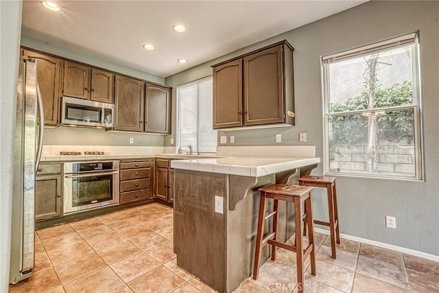 kitchen featuring stainless steel appliances, tile countertops, a peninsula, recessed lighting, and a breakfast bar