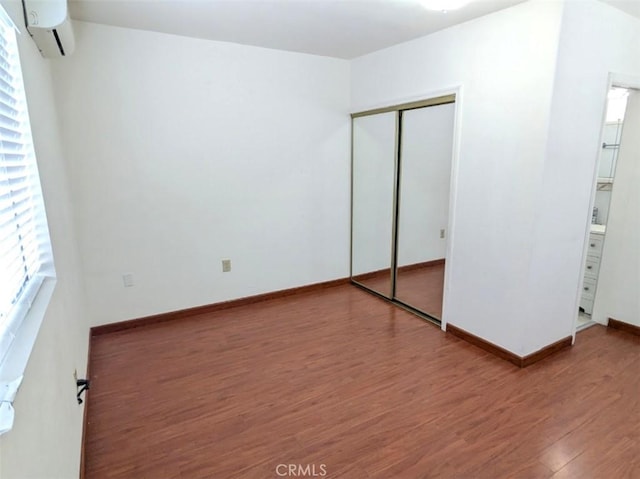 unfurnished bedroom with a wall unit AC, wood finished floors, a closet, baseboards, and connected bathroom