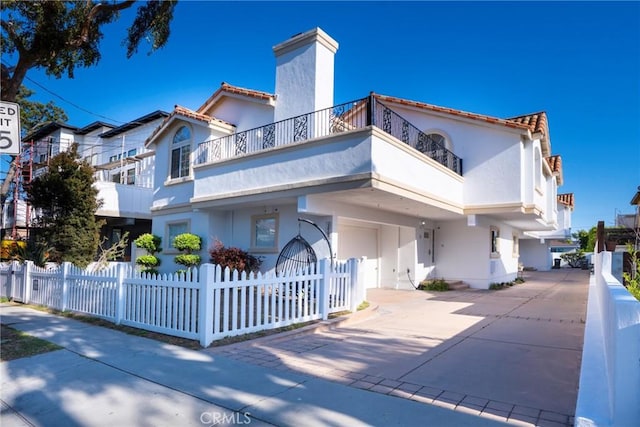 back of house featuring a balcony, a fenced front yard, stucco siding, a chimney, and concrete driveway