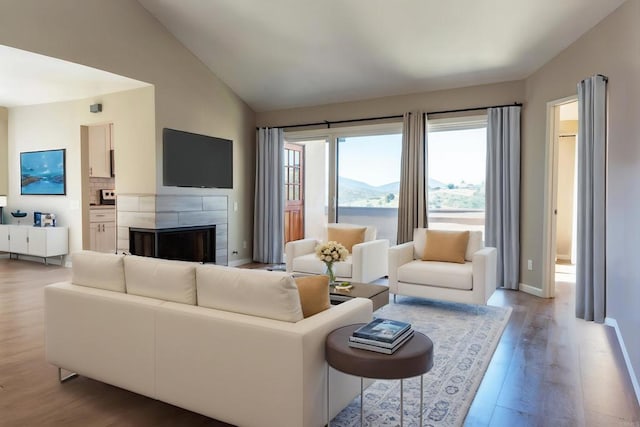 living area with wood finished floors, vaulted ceiling, a tiled fireplace, and baseboards