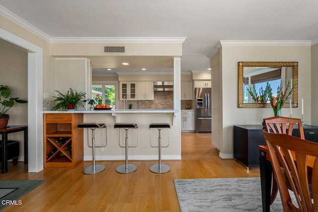 kitchen with stainless steel refrigerator with ice dispenser, a kitchen bar, light wood finished floors, decorative backsplash, and crown molding