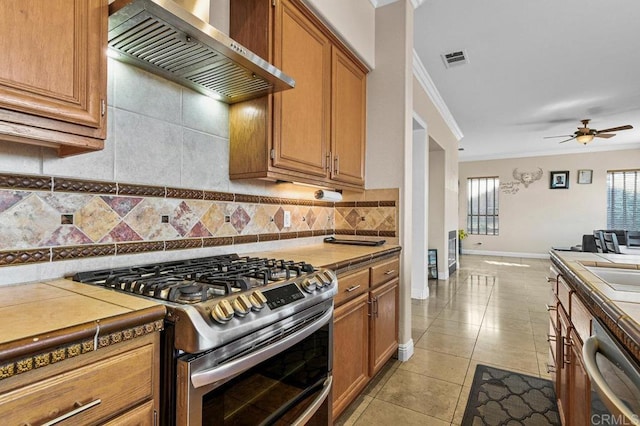 kitchen featuring stainless steel appliances, wall chimney exhaust hood, tile countertops, tasteful backsplash, and a ceiling fan