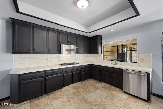 kitchen with appliances with stainless steel finishes, a sink, tile countertops, a raised ceiling, and backsplash