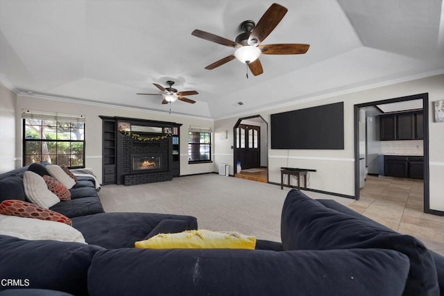 living area featuring a tray ceiling, ornamental molding, a brick fireplace, a ceiling fan, and baseboards