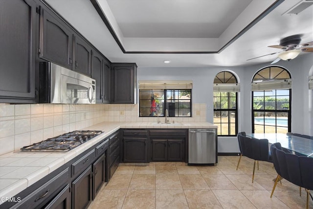 kitchen with tile counters, appliances with stainless steel finishes, a sink, a raised ceiling, and backsplash