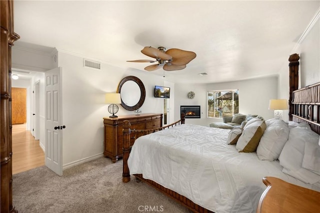 bedroom featuring light colored carpet, baseboards, ornamental molding, a glass covered fireplace, and ceiling fan