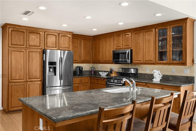 kitchen featuring appliances with stainless steel finishes, brown cabinetry, glass insert cabinets, dark stone counters, and light wood-type flooring