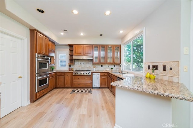 kitchen featuring appliances with stainless steel finishes, a peninsula, a sink, brown cabinets, and light stone counters