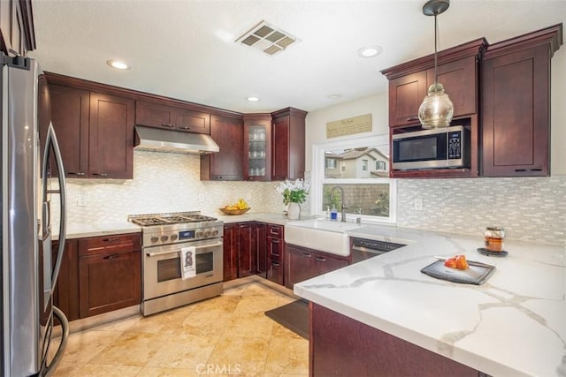 kitchen featuring stainless steel appliances, under cabinet range hood, a sink, light stone countertops, and decorative light fixtures