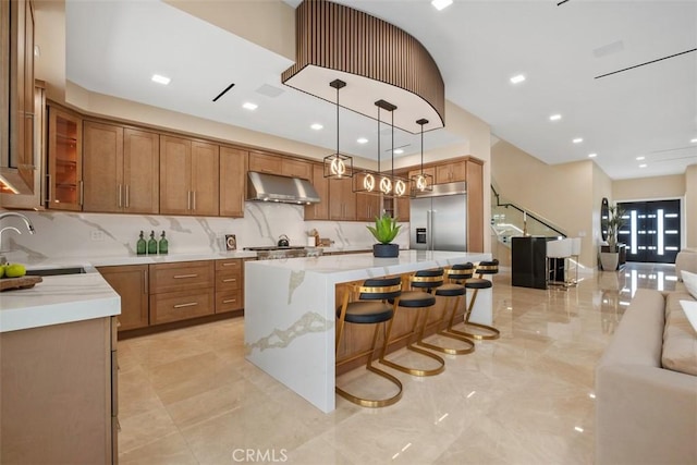 kitchen featuring appliances with stainless steel finishes, under cabinet range hood, open floor plan, a sink, and brown cabinetry