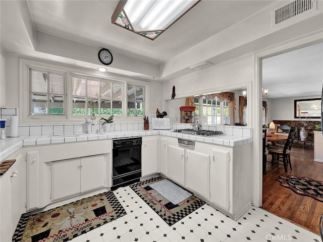 kitchen featuring black dishwasher, tile countertops, white microwave, a sink, and white cabinets