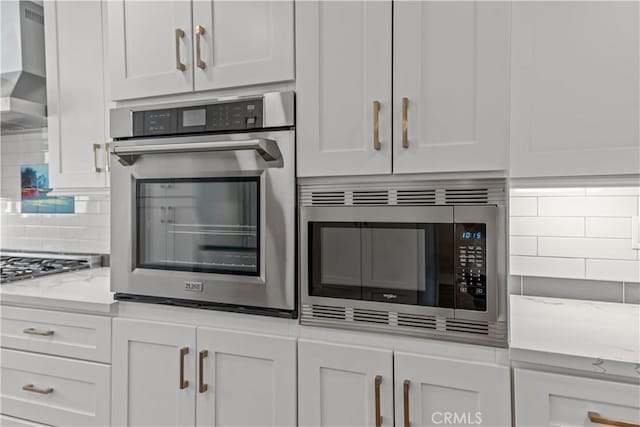 kitchen featuring appliances with stainless steel finishes, decorative backsplash, wall chimney range hood, white cabinetry, and light stone counters