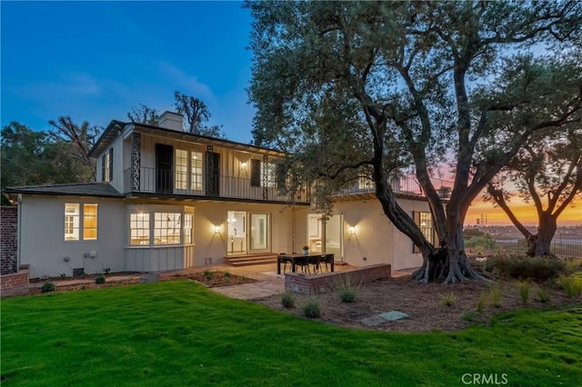 back of house with a balcony, a lawn, a chimney, stucco siding, and a patio