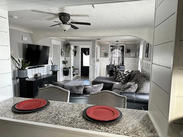 living area featuring arched walkways, a ceiling fan, a textured ceiling, and a wainscoted wall