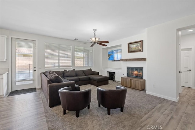 living room featuring light wood-type flooring, a ceiling fan, a glass covered fireplace, and baseboards